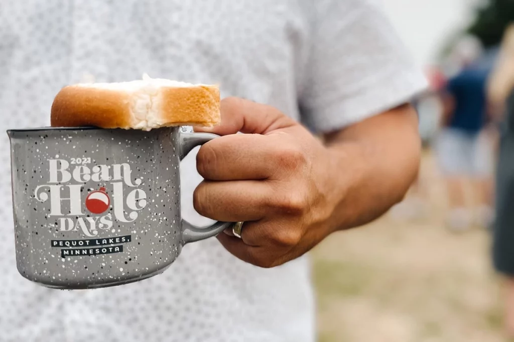 Horizontal Photo of Bean Hole Days showing a man holding a gray Bean Hole Days mug with a piece of bread on a summer day