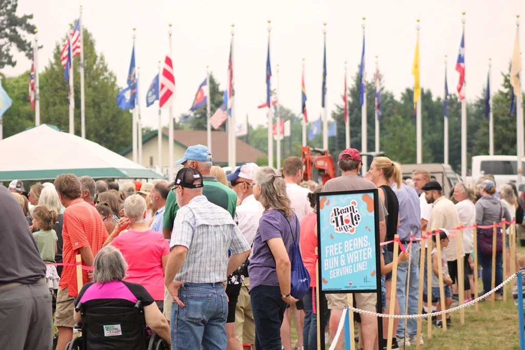 Photo of Bean Hole Days showing a crowd of people lined up to get beans with flags and trees in the background on a summer day