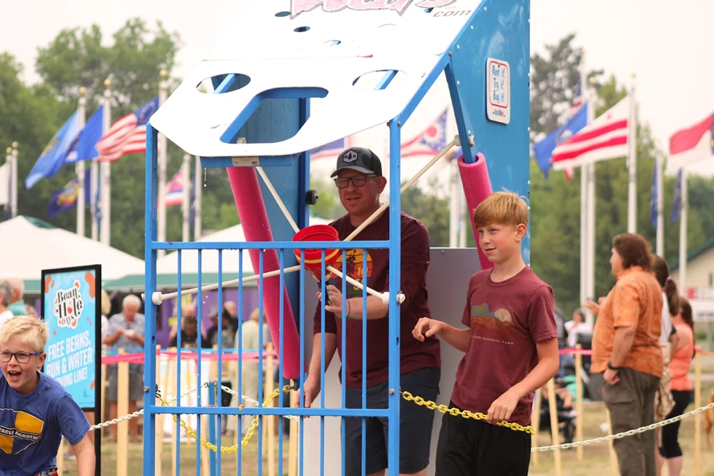 Photo of Bean Hole Days showing a dad with two sons playing with water balloons at a fair game with crowds of people in the background on a summer day