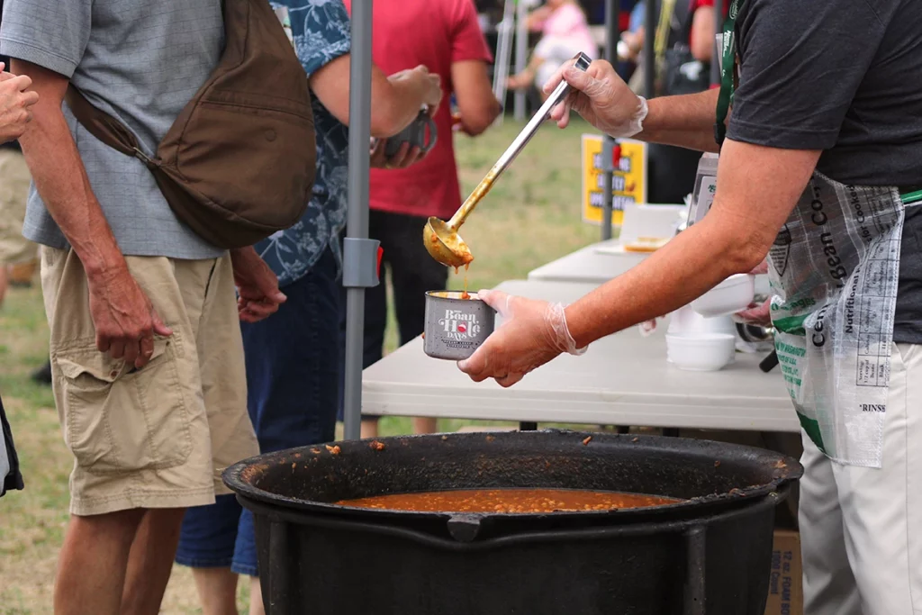 Photo of Bean Hole Days showing a volunteer scopping beans out of a huge iron kettle into a mug and handing it to a man in line on a summer day