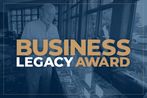 Photo of an older man Bob Spizzo in a large room at Breezy Point Resort next to a large collection of old newspapers with a blue overlay and a tan and white Business Legacy Award heading on top