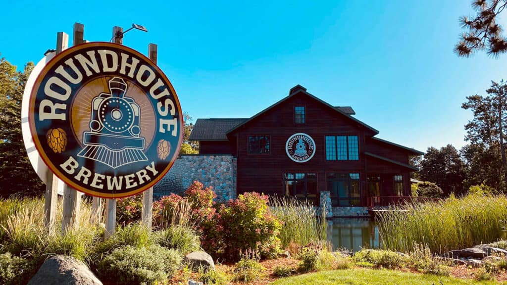 Photo of the exterior of a large cabin like building Roundhouse Brewery on a sunny summer day showing landscaping and a large logo sign