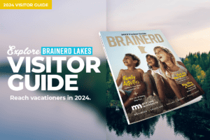 Photo of the 2023 Brainerd Lakes Visitor Guide magazine showing the cover with a photo of smiling children at a beach on top of an aerial photo of a calm lake with forest in the background at sunset