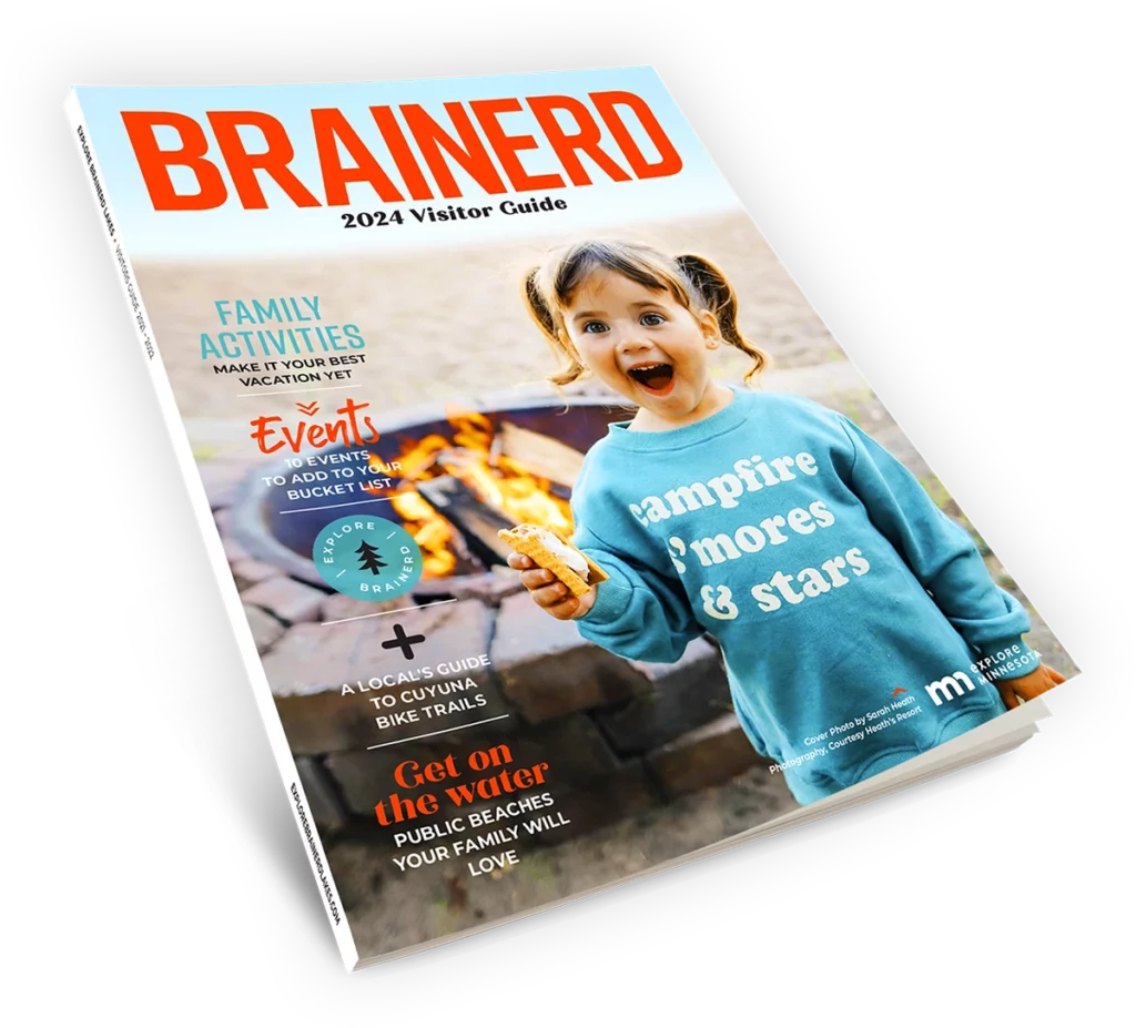 A 2024 Brainerd Lakes Visitor Guide magazine showing the cover with a large photo of a smiling little girl wearing a blue seatshirt and eating a smore next to a campfire